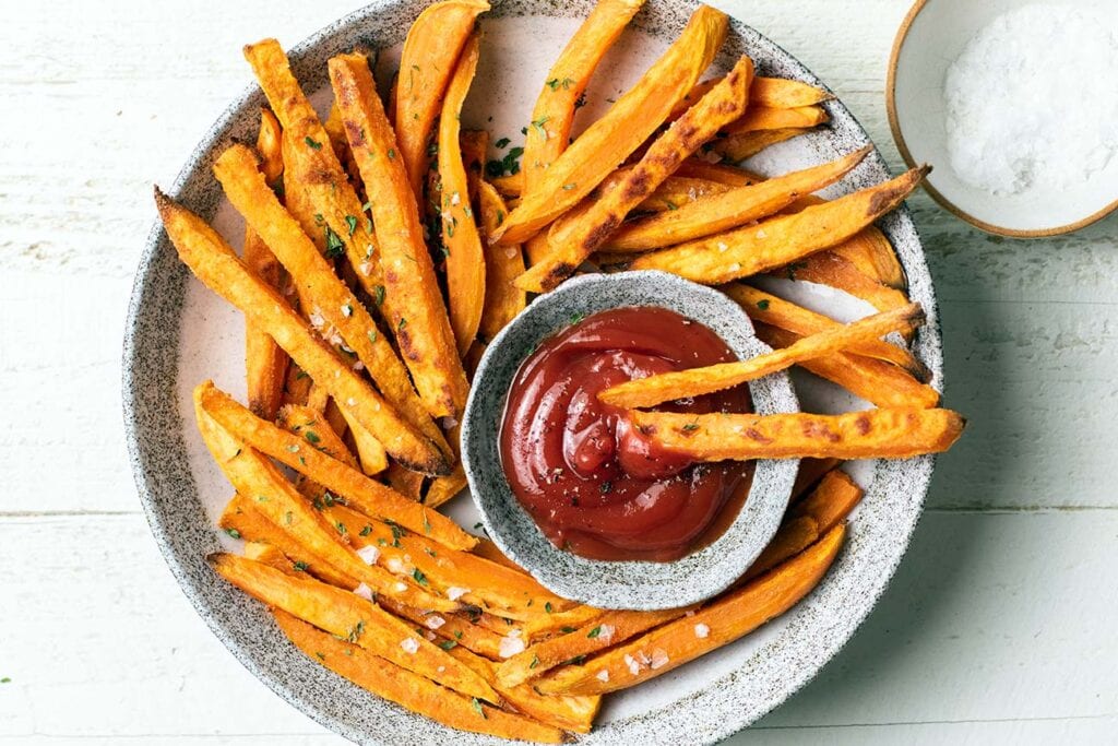A plate with crisp baked sweet potato fries surrounding a dish of ketchup.