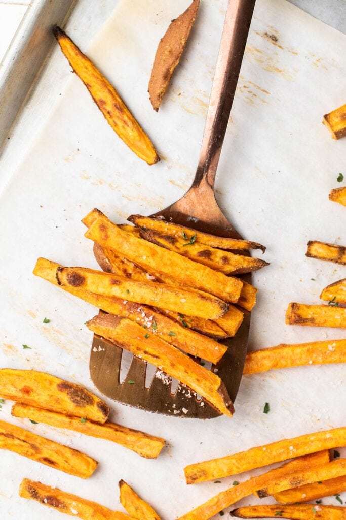 A spatula picking up browned crispy sweet potato fries off a lined baking sheet.