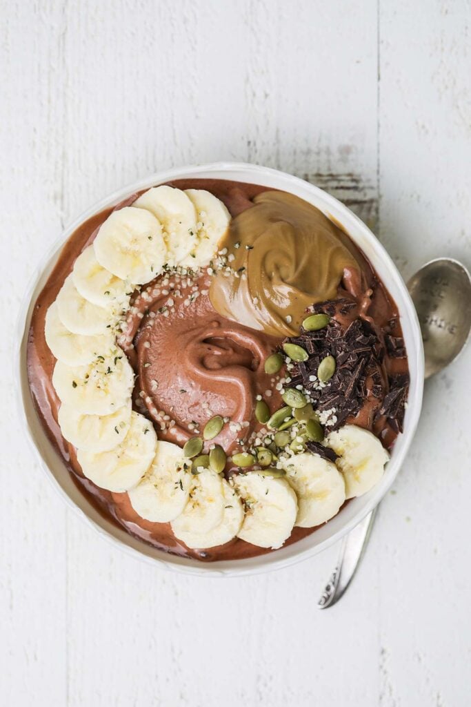 A chocolate smoothie in a bowl topped with banana, nut butter, and seeds.