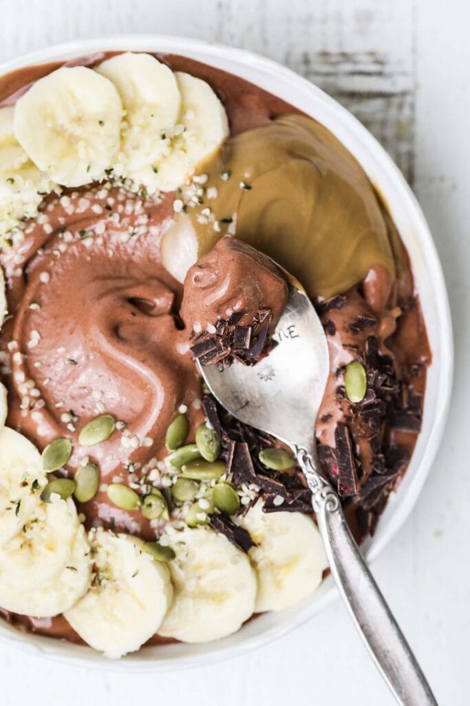 A spoon taking a bite of a thick chocolate smoothie bowl garnished with banana and seeds.