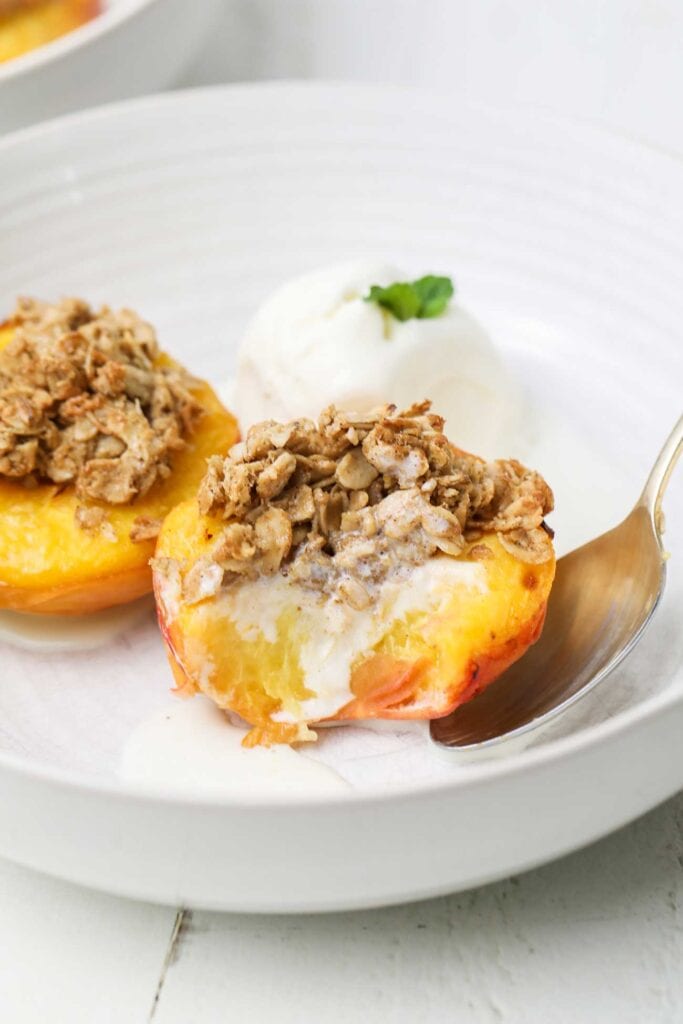 A bowl with two peaches cooked with an oatmeal crisp topping with ice cream.