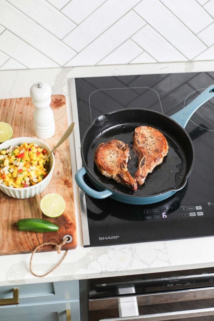 Pork chops shown in a cast iron skillet cooking on a Sharp induction cooktop.