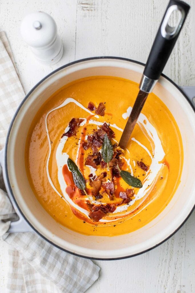 A pot of Butternut Squash Red Pepper Soup shown garnished with bacon, sage, and hot sauce.