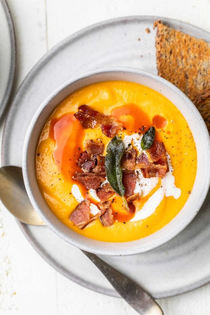 A bowl or butternut squash red pepper soup garnished with bacon, sage, and hot sauce.