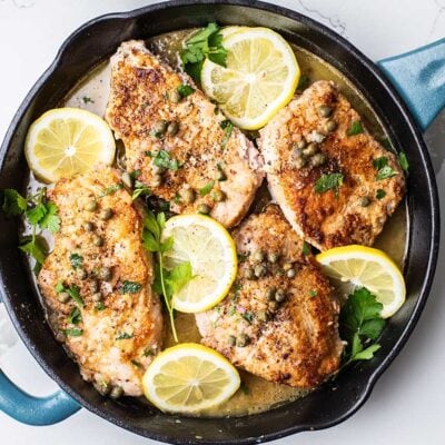 A skillet with the pork chops added back in, garnished with lemon slices, parsley, and capers.