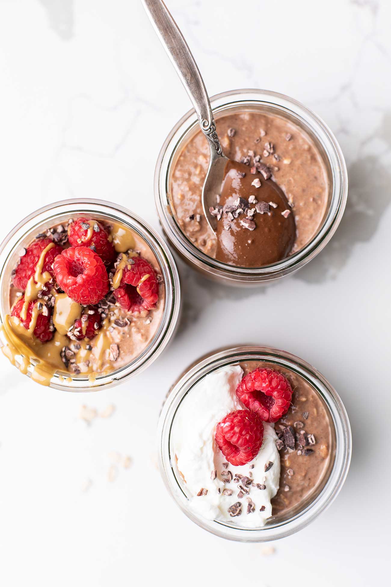 Three jars of overnight oats topped with raspberries, yogurt, and cacao nibs.