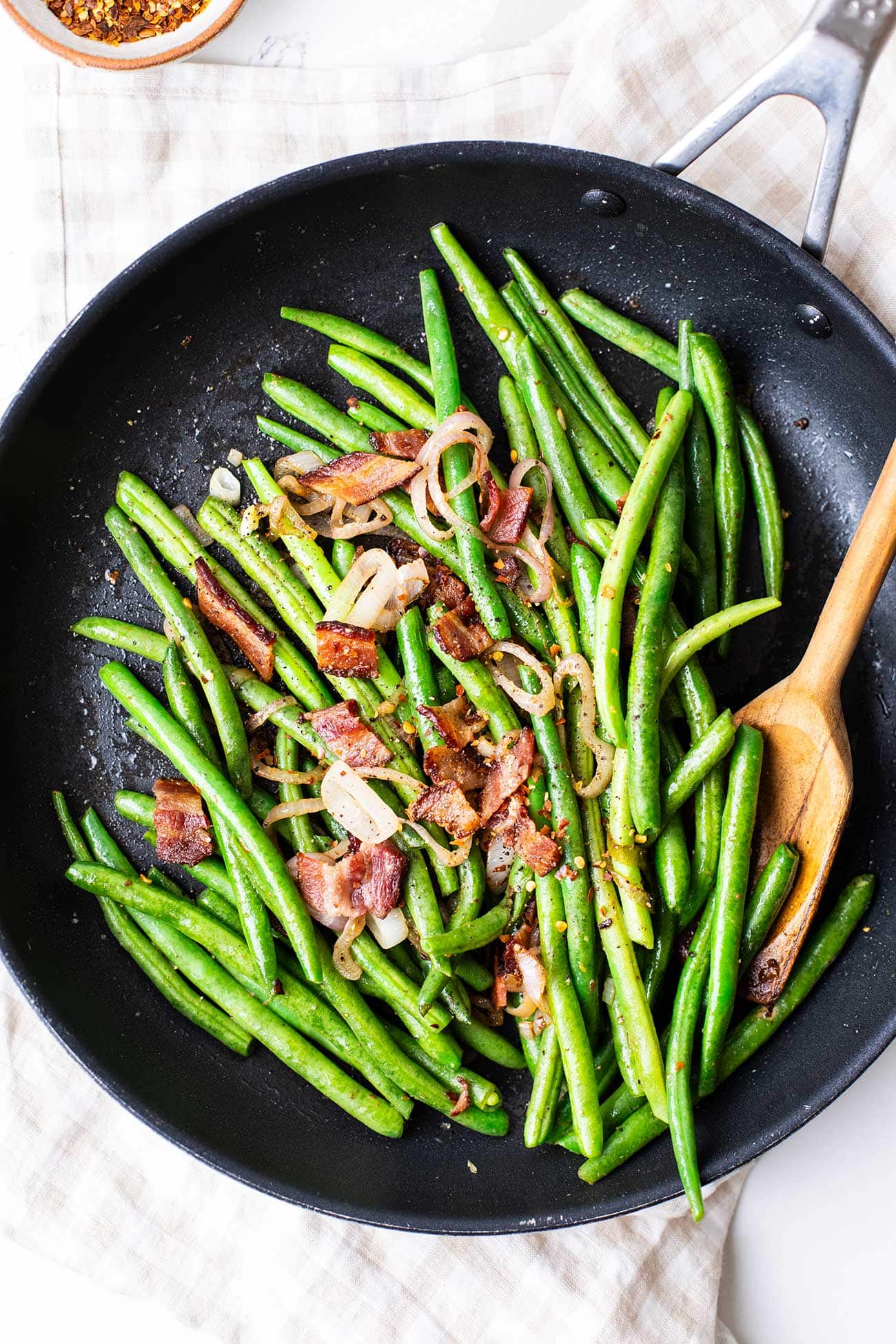 A skillet filled with bacon green beans with a wooden spoon.