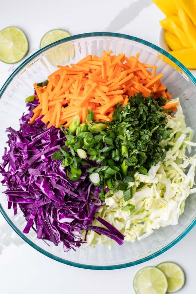 A large glass bowl shown with shredded cabbage, shredded carrots, and fresh chopped herbs.