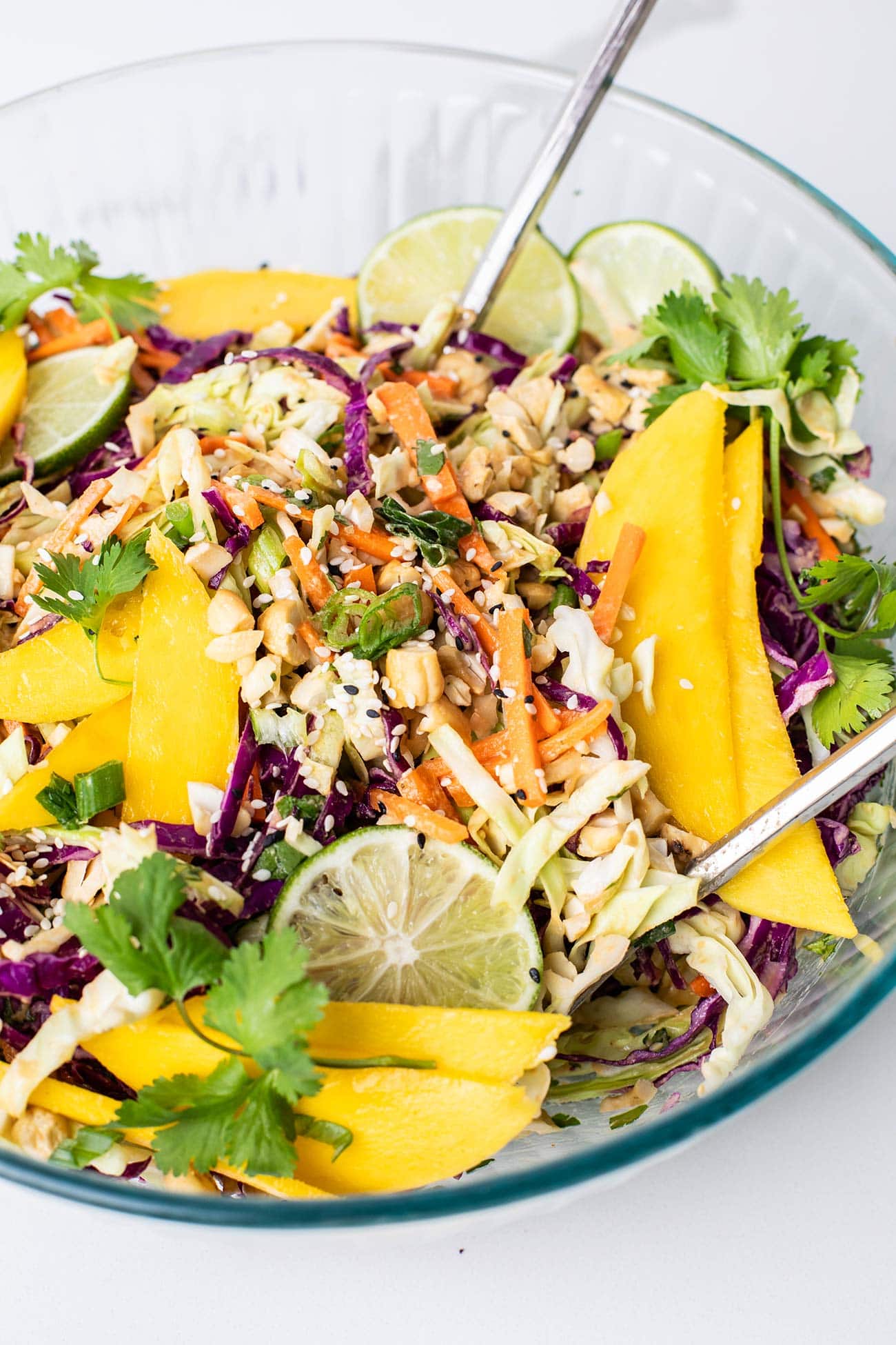 A glass bowl with a colorful cabbage and carrot salad topped with mango and cashews.