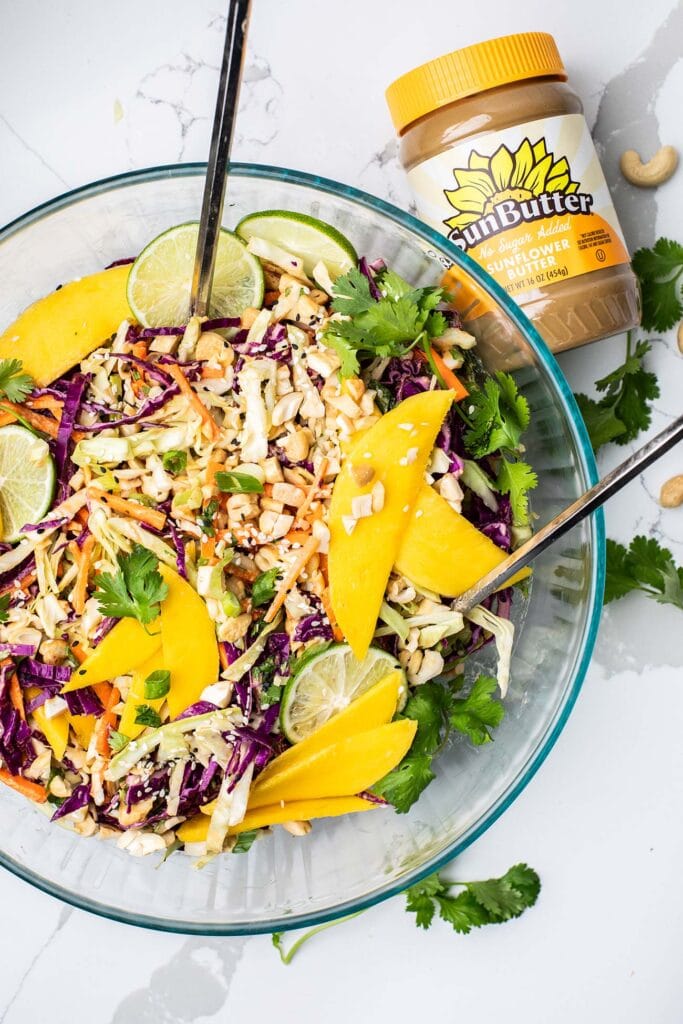 A large bowl showing a carrot and cabbage salad topped with mango next to a jar of SunButter sunflower seed butter.