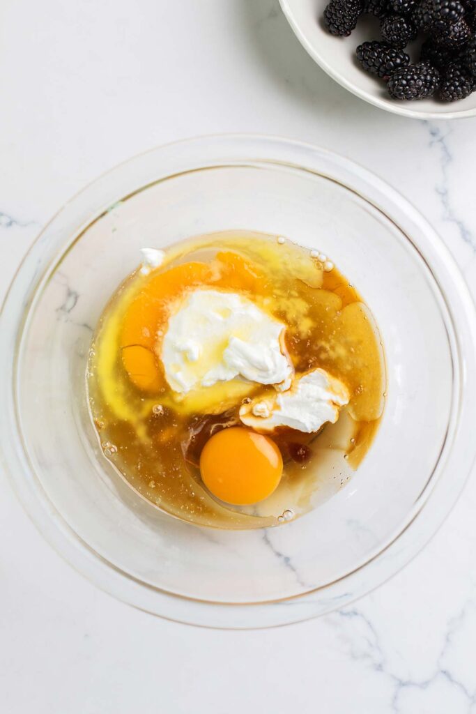 A glass bowl shown with eggs, oil, yogurt, and vanilla.