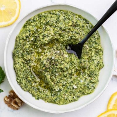 A bowl of vibrant green basil walnut pesto shown with lemon slices and garlic cloves next to the bowl.