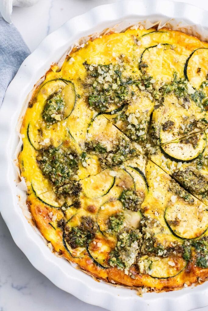 A zucchini quiche topped with lots of basil pesto shown cut into wedges to serve.
