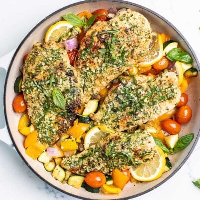 4 chicken breasts coated with pesto sitting in a skillet with colorful vegetables.