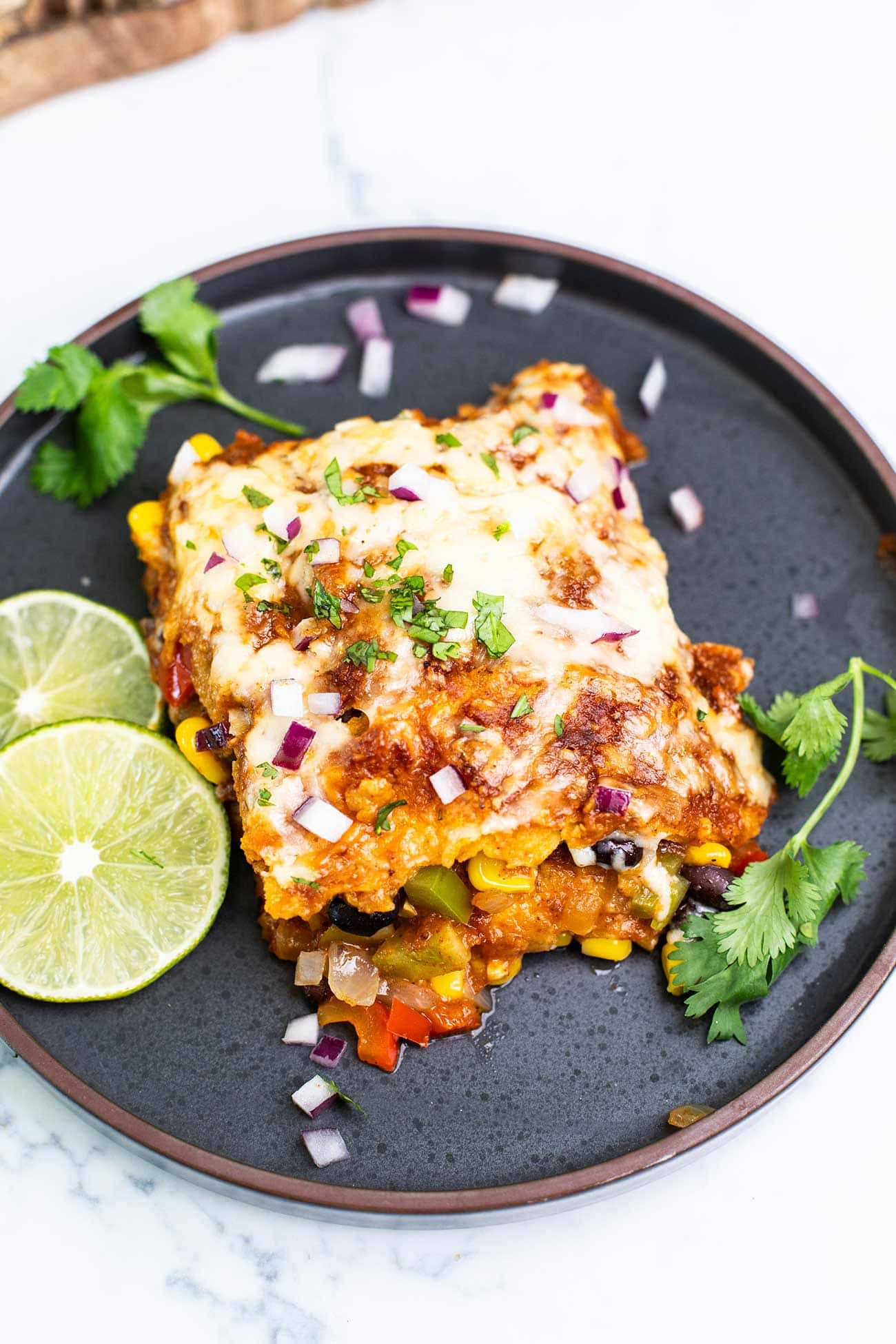 A slice of enchilada casserole on a black plate garnished with onions and lime slices.