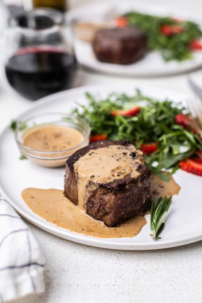 A filet mignon drizzled with peppercorn sauce, with an extra ramekin of sauce next to the steak.