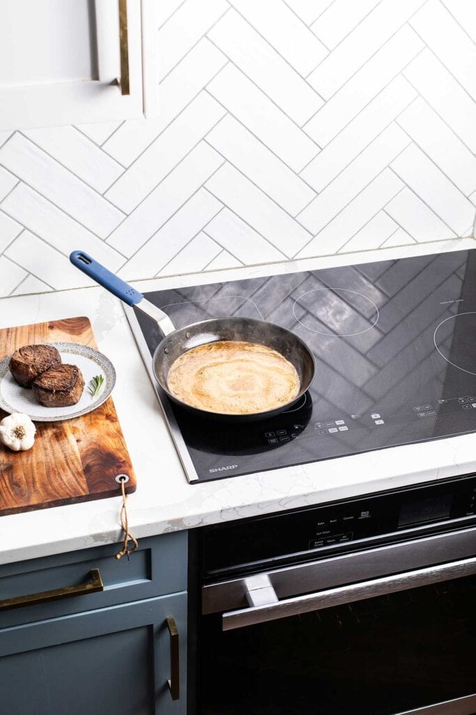 A creamy peppercorn sauce simmering to thicken next to steaks resting on a plate.