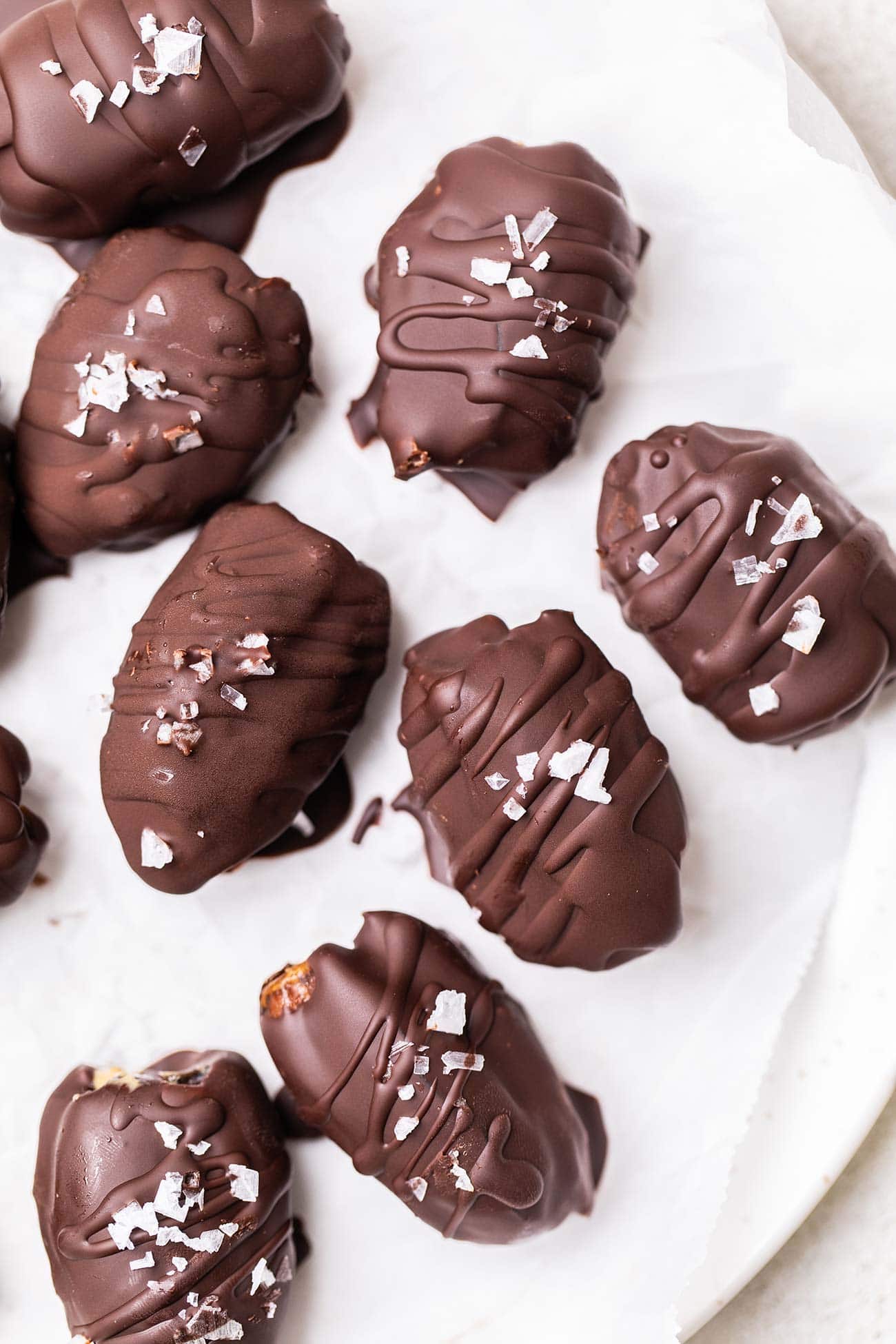 A white plate with dark chocolate covered dates sprinkled with flaky salt.