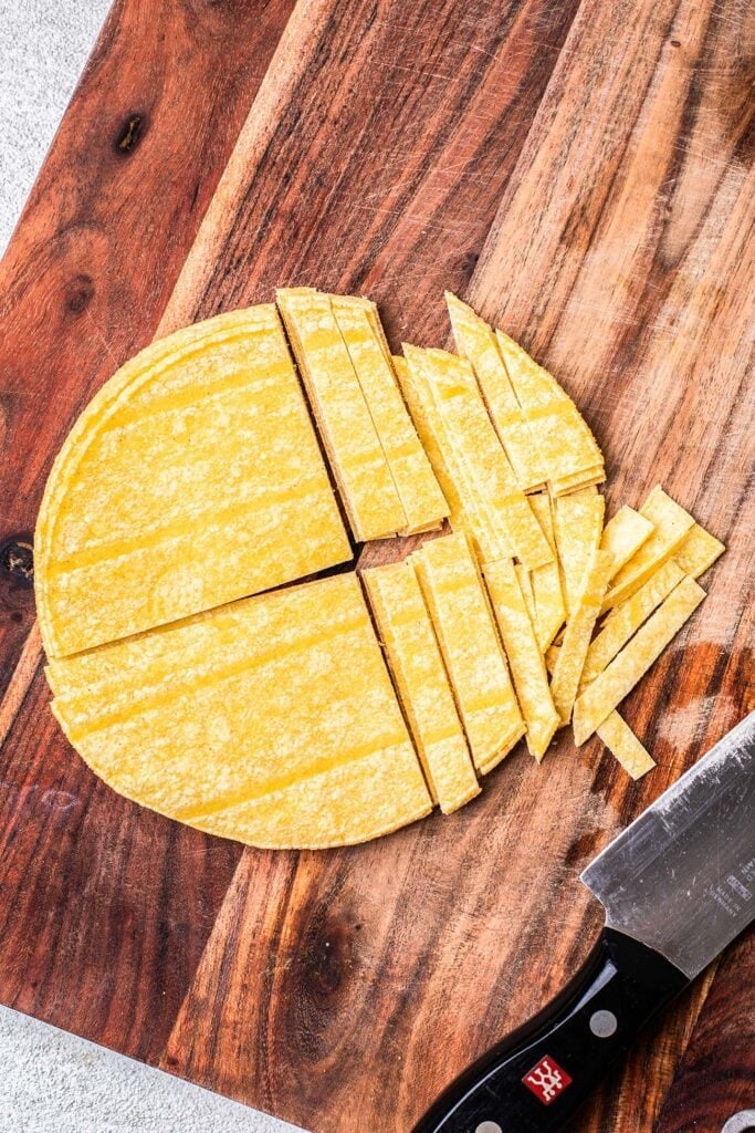 Corn tortillas on a cutting board being cut into thin strips.