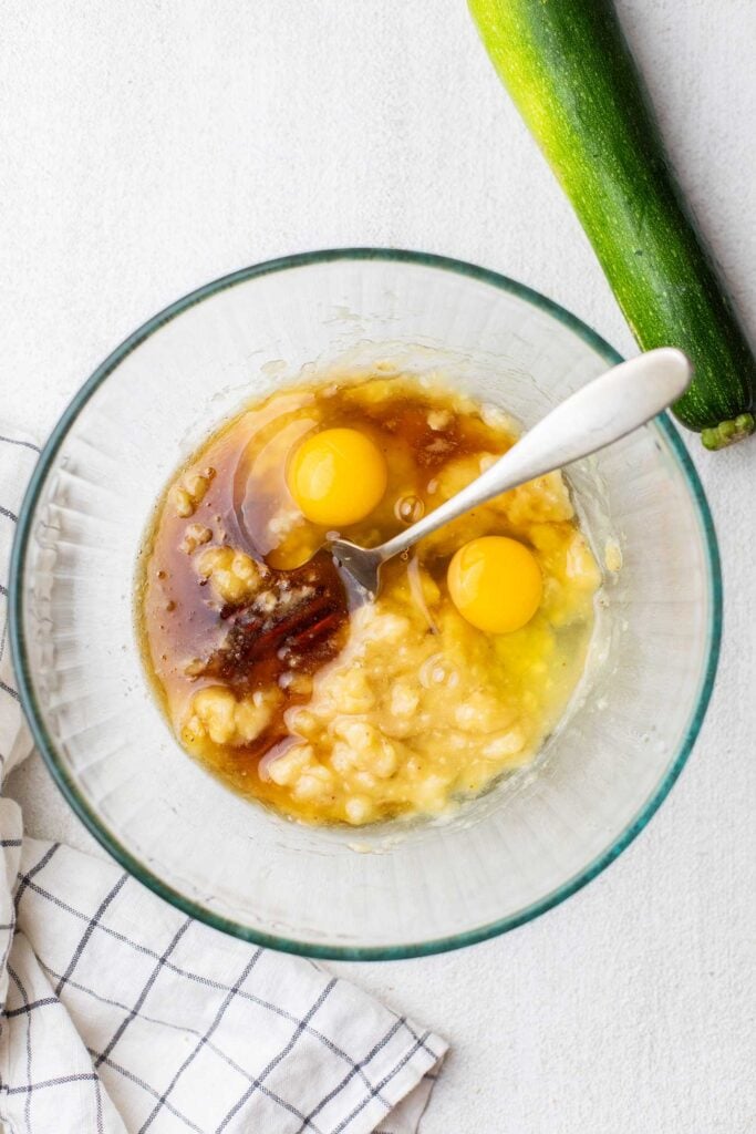 Eggs, coconut oil, and vanilla added to the mashed banana in a mixing bowl.