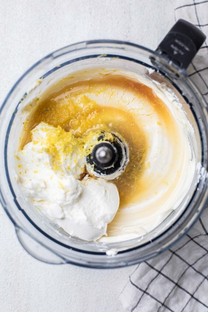 Cream cheese in a food processor bowl with yogurt, lemon juice, lemon zest, and maple syrup added in.