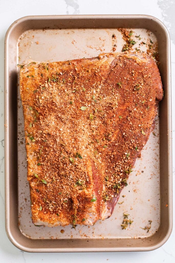 A brisket on a baking pan shown rubbed with spice rub and covered with plastic wrap.
