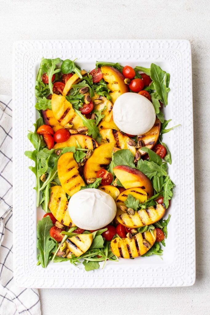A platter of grilled peach salad showing adding the burrata cheese and pistachios.
