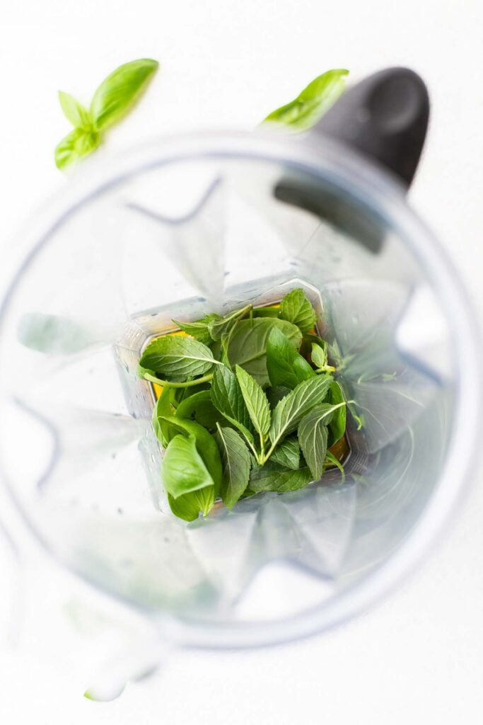 A blender with olive oil and fresh herbs shown inside.