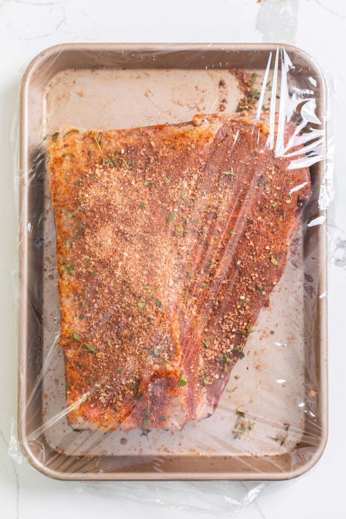 A brisket coated in spices on a baking tray wrapped in plastic wrap.
