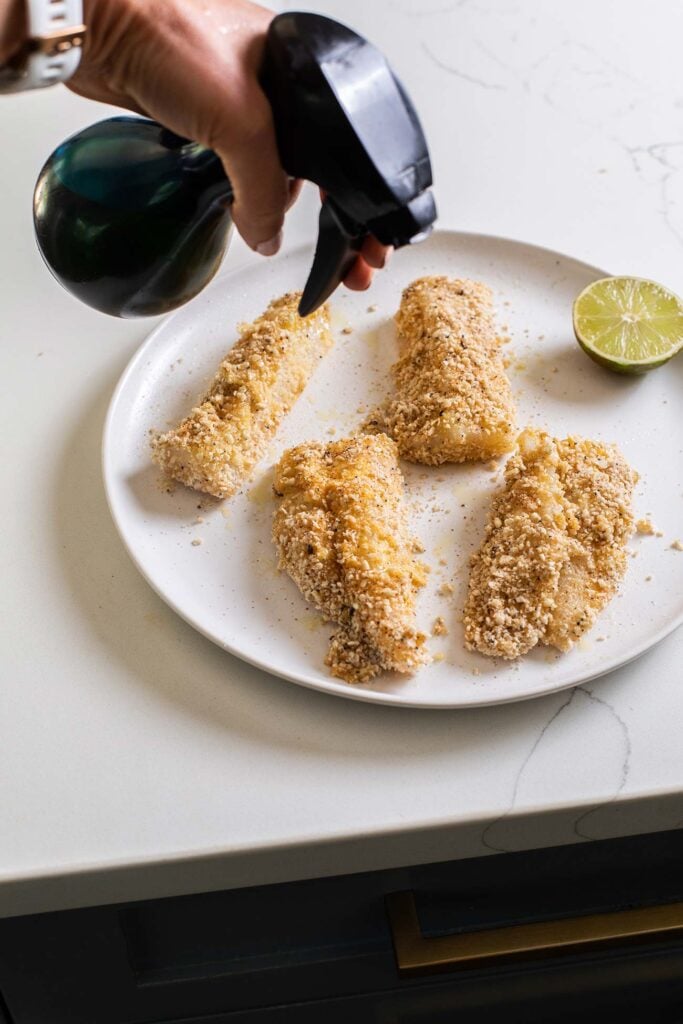 An oil sprayer spraying the breadcrumb coated pieces of fish.