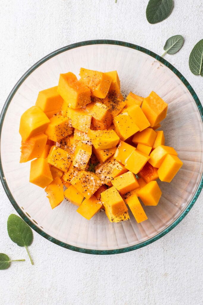 A bowl of raw butternut squash cut into cubes, seasoned with olive oil, salt and pepper.
