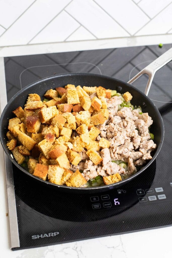 Cornbread and cooked sausage added to a skillet with softened vegetables.