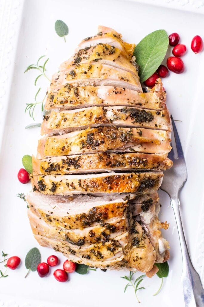 A turkey breast on a white platter shown garnished with herbs and cranberries.