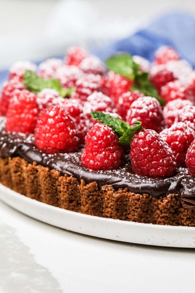 A raspberry chocolate tart shown garnished with fresh raspberries, mint leaves, and powdered sugar.