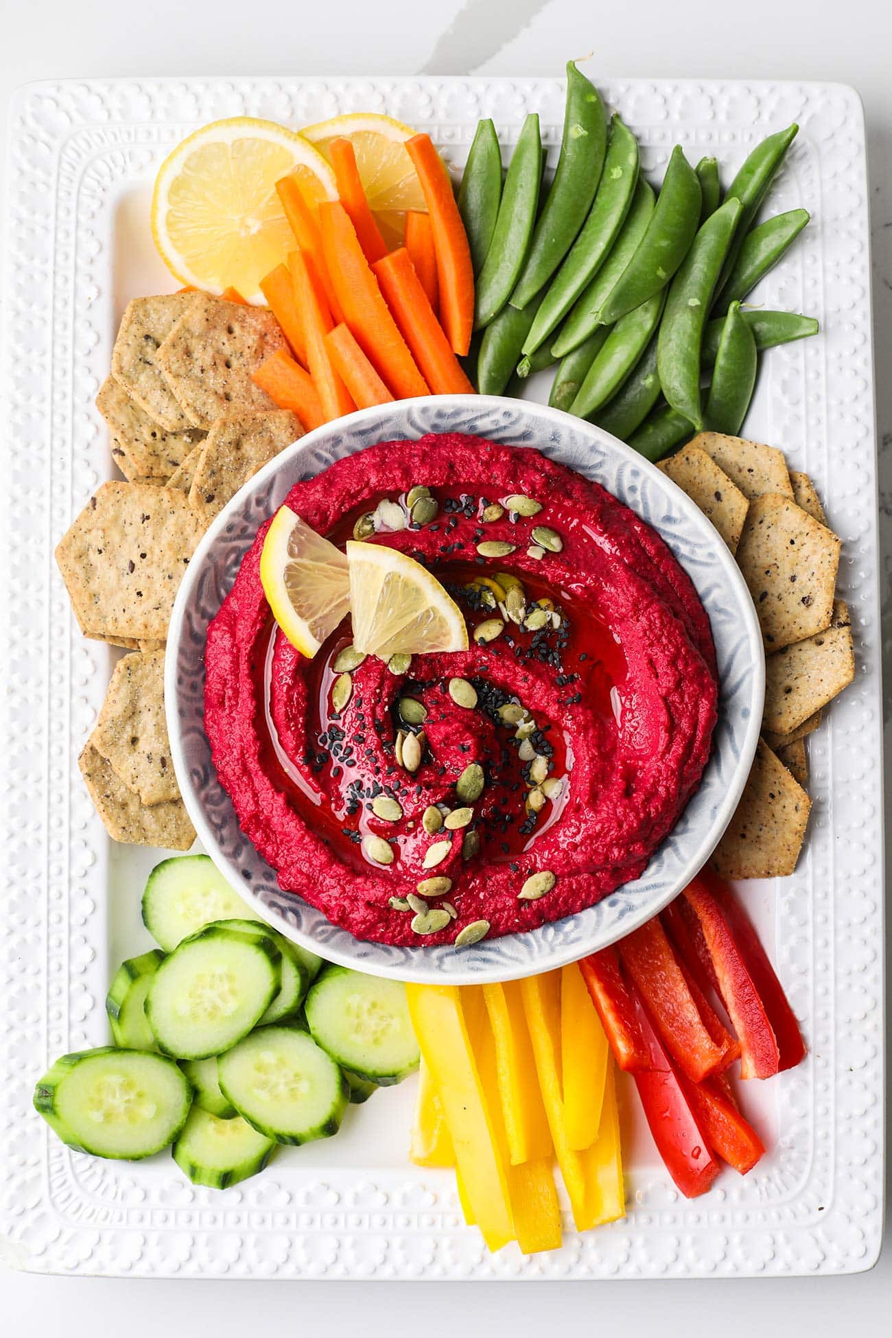 A bowl of pink hummus on a platter with colorful veggies and crackers.