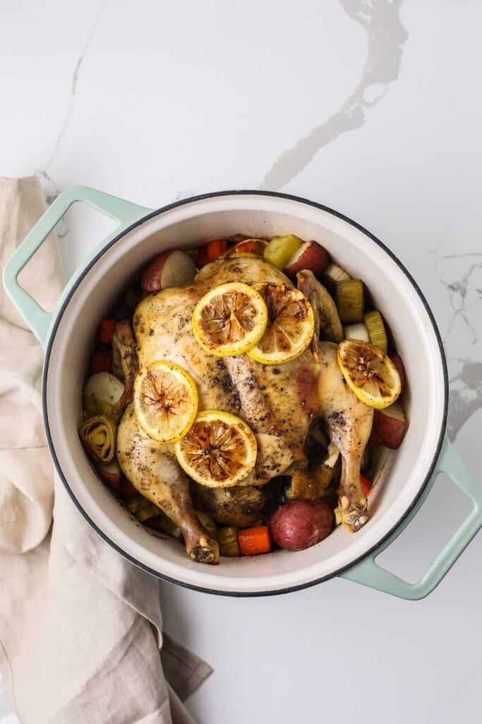 A whole roasted chicken with lemon slices surrounded by vegetables in a dutch oven.