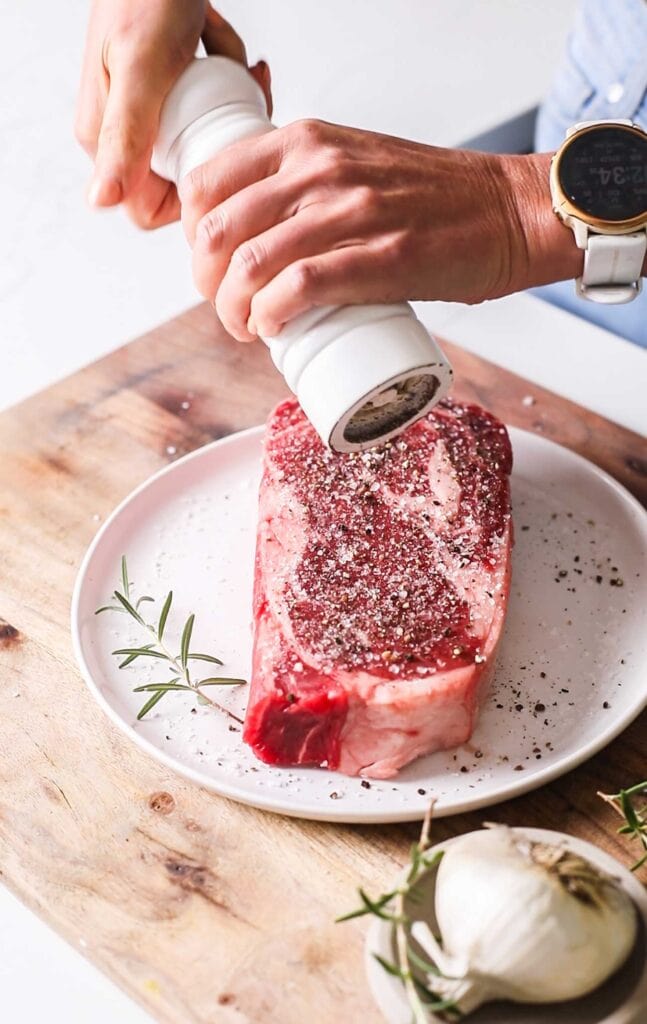 A steak being generously seasoned with Kosher salt and black pepper.
