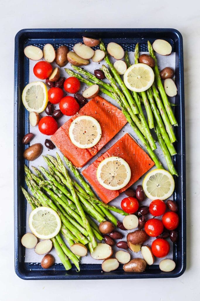 A sheet pan with salmon and asparagus, along with baby potatoes, olives, and tomatoes, seasoned and garnished with lemon slices.