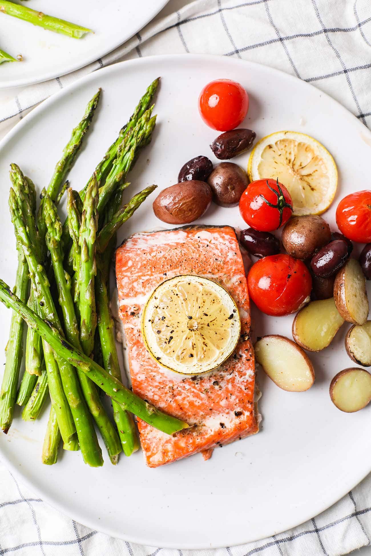 A white plate with a salmon and asparagus dinner, including baby potatoes, tomatoes, and olives.