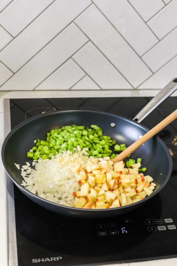 A skillet on the stovetop with onions, celery, chopped apples, and garlic.
